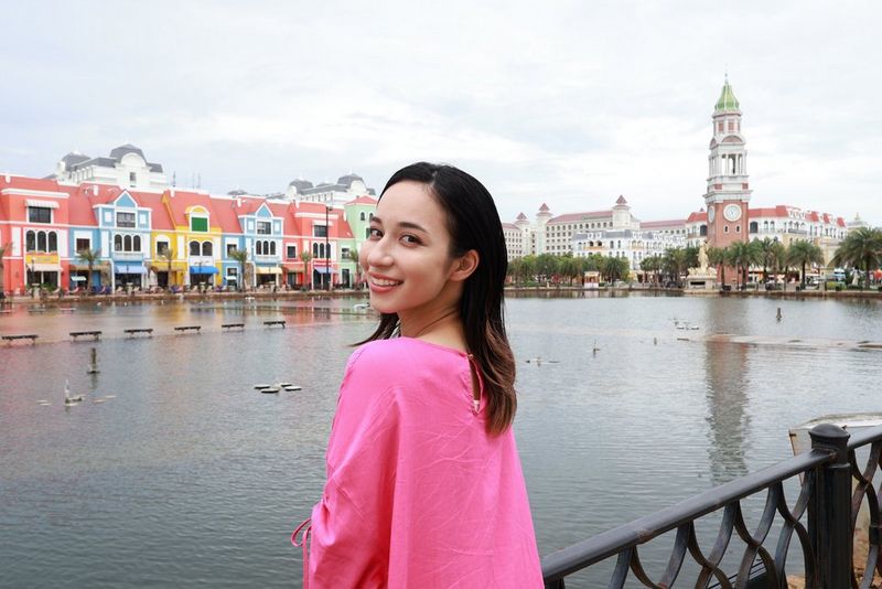 Повернувши голову, щоб посміхнутися в камеру, молода жінка стоїть на березі річки, а навпроти неї — кілька барвистих будинків і більші будівлі вдалині. Знято за допомогою ширококутного об’єктива Canon RF-S 10-18mm F4.5-6.3 IS STM.