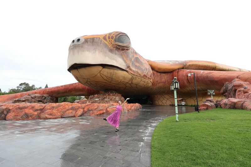 Une femme vêtue d'une robe rose vif pose sur une chaussée mouillée devant un bâtiment en forme de tortue géante. Photo prise avec un objectif à grand-angle Canon RF-S 10-18mm F4.5-6.3 IS STM.