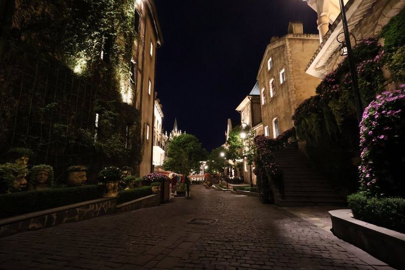 Ноќна улична сцена со улица со калдрма, камени згради и градина со скулптури во форма на глави со зеленило наместо коса, сето многу детално. Фотографирано со објектив Canon RF-S 10-18mm F4.5-6.3 IS STM.
