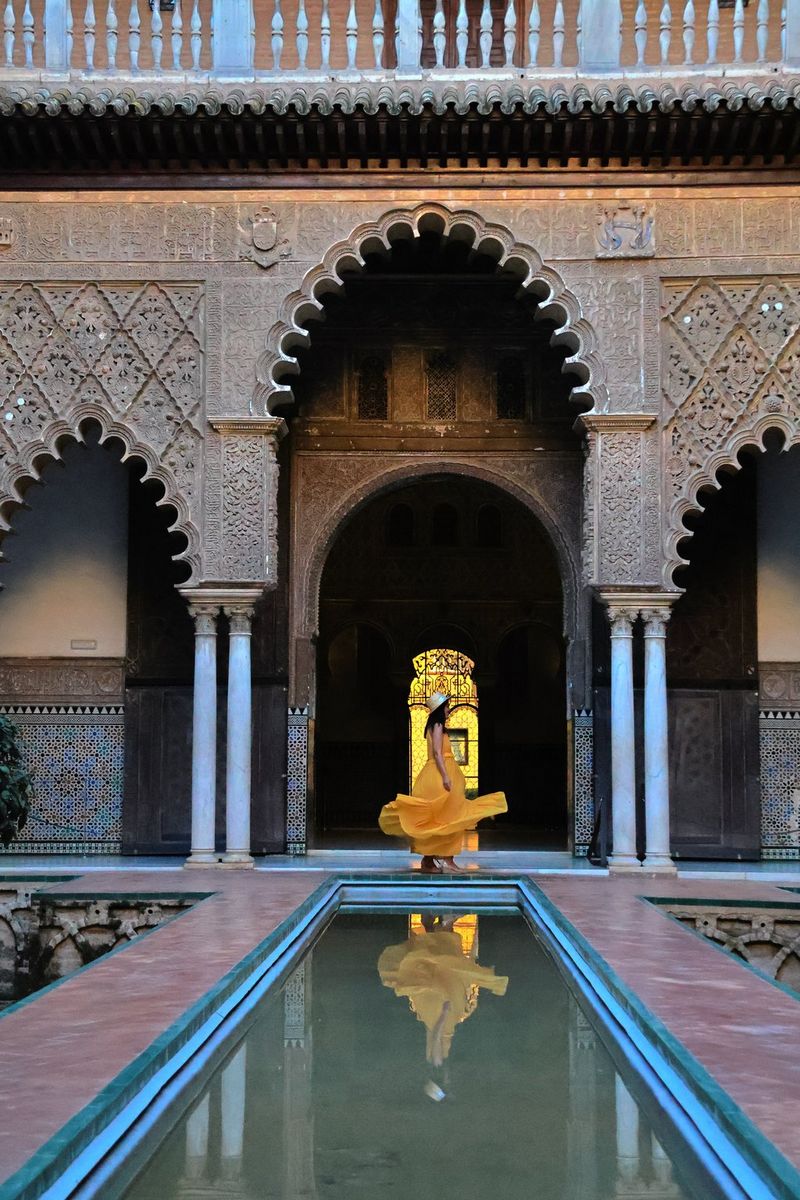 A woman in a yellow dress stands beneath an ornate archway in front of a long, narrow pool of water in which her reflection can be seen. Taken with a Canon RF-S 18-150mm F3.5-6.3 IS STM lens.