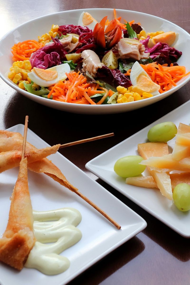 A picture of a salad bowl filled with shredded carrots, boiled egg quarters, sliced red cabbage, mushrooms and sweetcorn. Two square plates in the foreground hold other foods.
