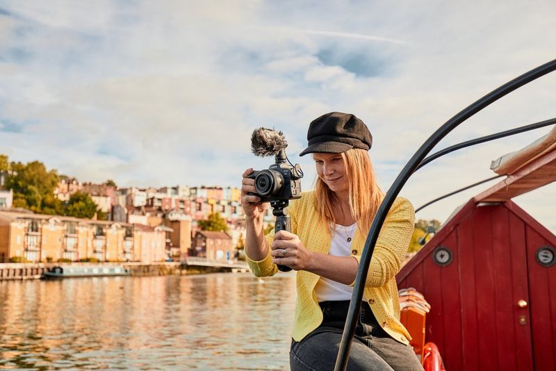 A young woman perched on the edge of a boat holding a Canon EOS R6 on a tripod grip, with a river and a row of houses in the background.