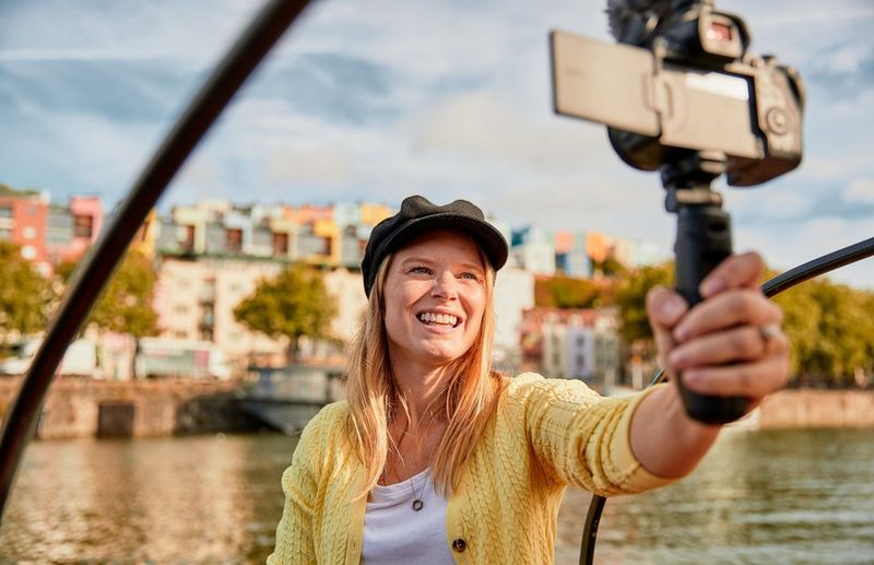Una donna punta verso sé stessa una fotocamera montata su un treppiede. Sullo sfondo, sfocata, una schiera di edifici multicolore sulla riva di un fiume.