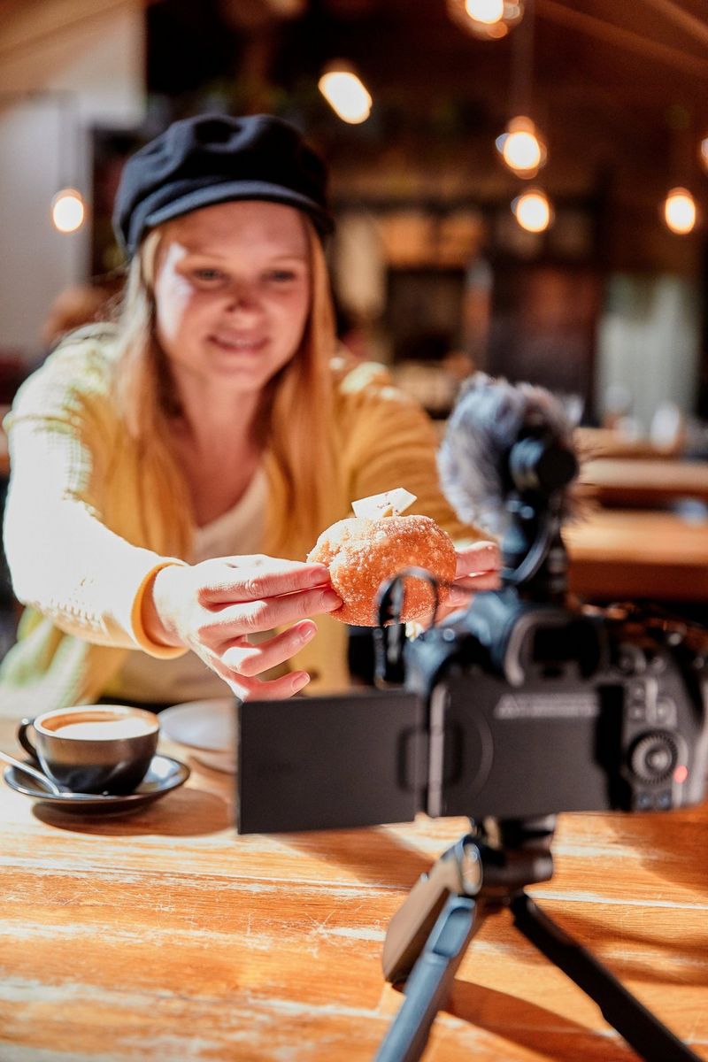 Женщина в кафе демонстрирует круассан на камеру, установленную на небольшом штативе на столе перед ней.
