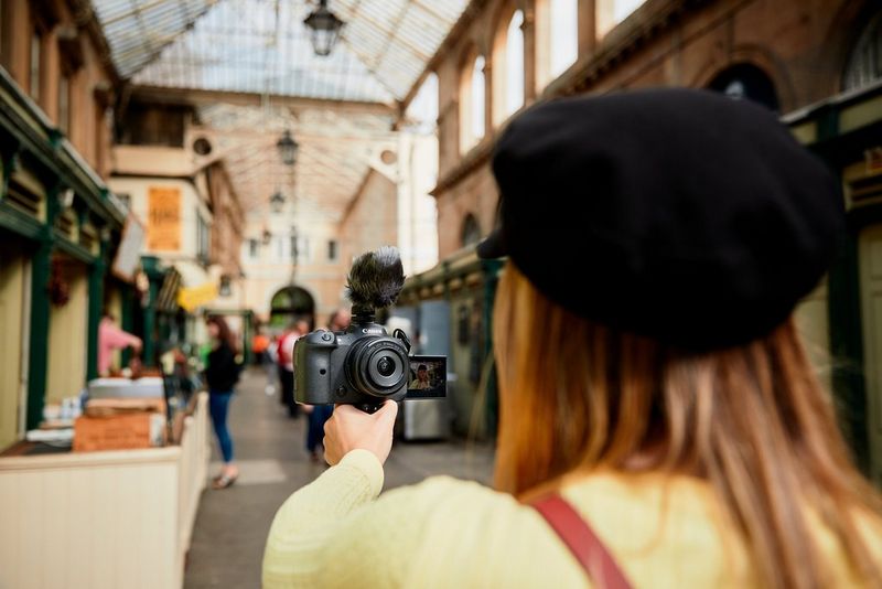 An over-the-shoulder shot of a young woman walking through a covered market, filming herself on a Canon EOS R6 camera.
