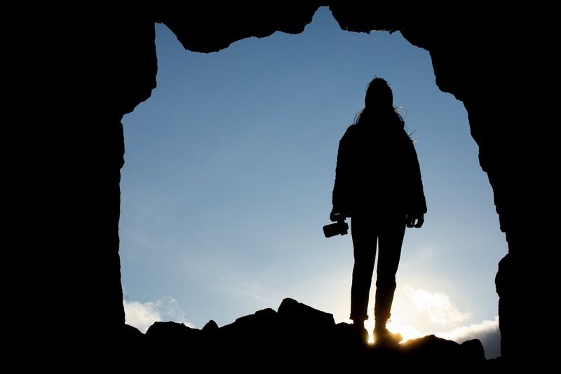A figure standing at the entrance to a cave holding a camera, silhouetted against a bright, sunny sky. 