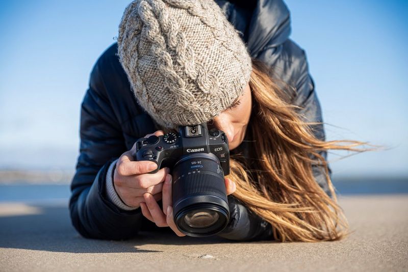 A woman takes a macro image of a shell with a Canon camera and lens. She is dressed in winter clothing and resting on her elbows in the sand for support.