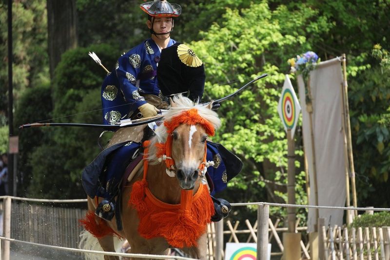 Een yabusame-boogschutter in ceremoniële kleding en met een grote pijl en boog die op hoge snelheid op een paard over een smal parcours rijdt.
