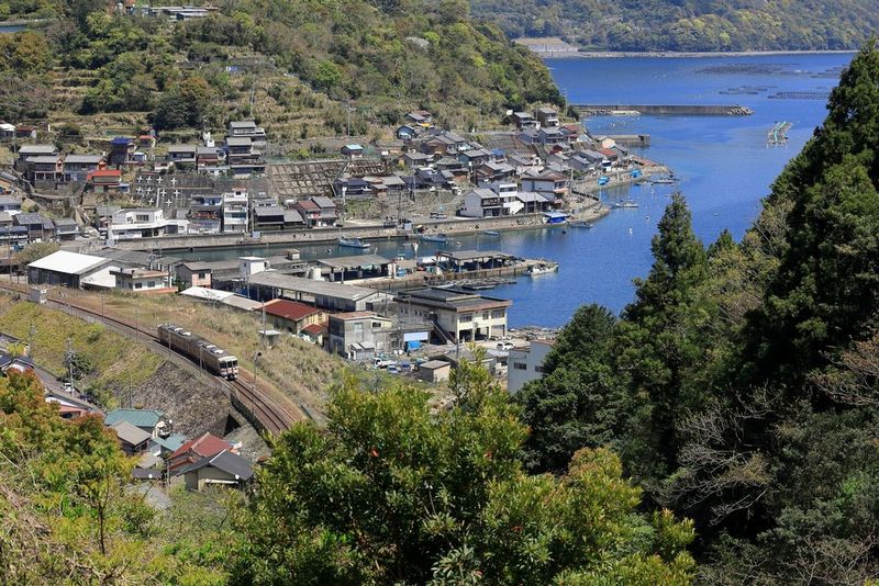 A view of a train travelling around an inlet with buildings on the hilly shore. The image is sharp from the foreground trees to the distant shoreline in the background.