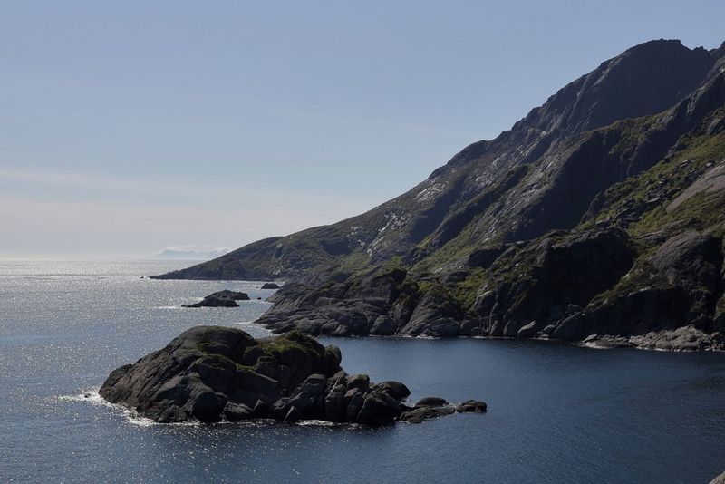 In un paesaggio costiero fotografato con un obiettivo Canon RF 24-240mm F4-6.3 IS USM, il mare risplende alla luce del sole intorno a un affioramento roccioso, con scogliere aspre che si innalzano dall'acqua dietro.