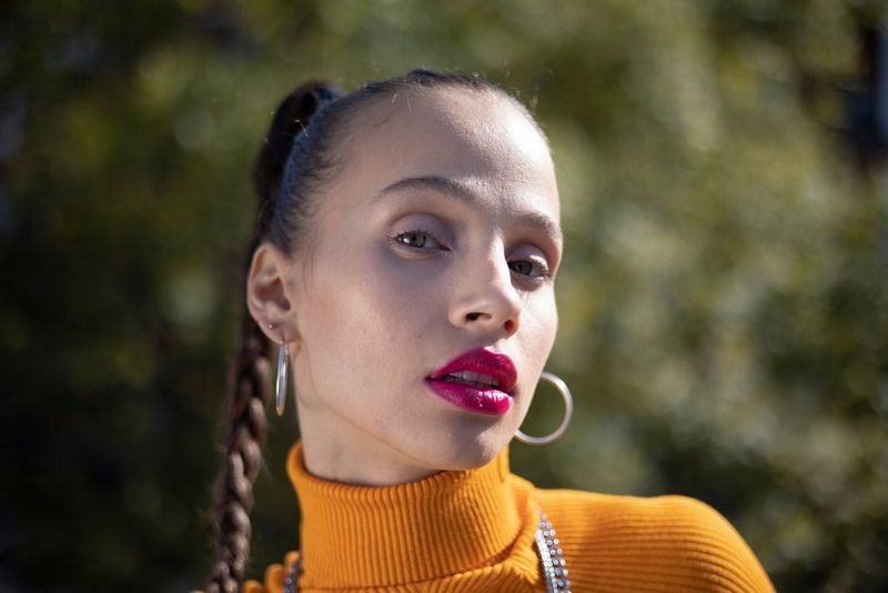 A portrait of a woman with bright pink lipstick and her hair scraped back in a plait against a blurred background. 