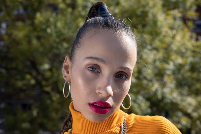 Портрет женщины с яркой розовой помадой в топе горчичного цвета на фоне размытого заднего плана, созданного с помощью широкой диафрагмы.