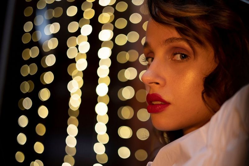 Женщина в белой блузке с жабо поворачивается к камере. Огни на заднем плане создают эффект боке.