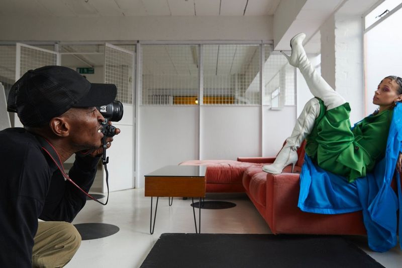 Фотограф Эджиро Дафе снимает женщину в белых ботинках и зеленом костюме на диване.