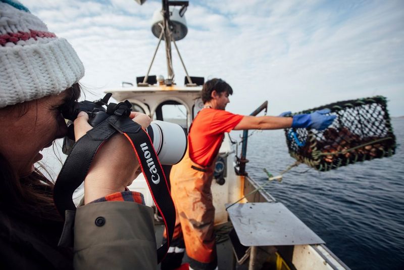 Een vrouw houdt een Canon EOS R System-camera bij haar hoofd en fotografeert een visser die een kreeftenval in de zee werpt.