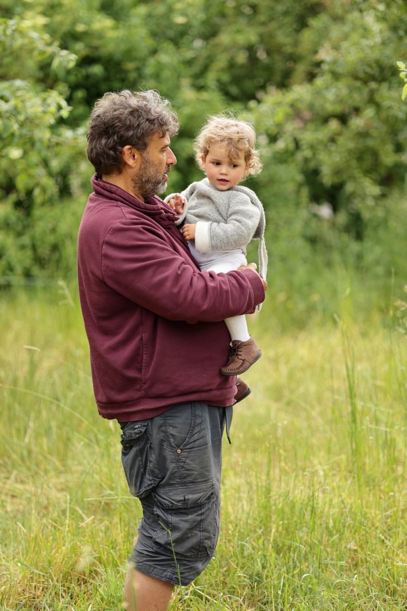 Osoba s bradom stoji u visokoj travi, držeći malo dijete.