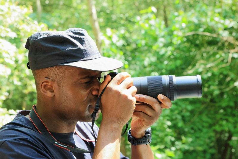 A photographer holding a Canon telephoto zoom lens for bird photography in a wooded area.