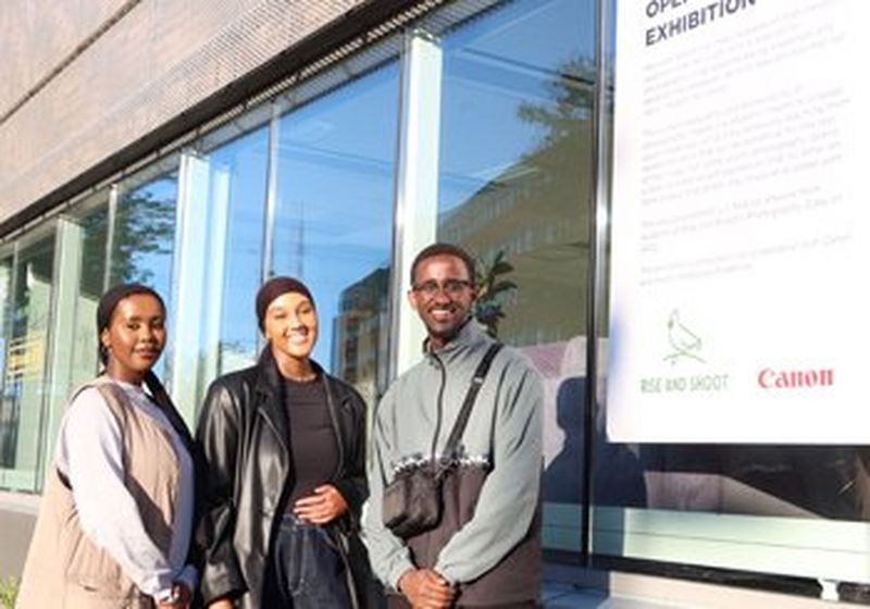 Three smiling young people stand beside a poster announcing the opening of an exhibition. The logos of Canon and Rise and Shoot can be clearly seen at the bottom of the poster.