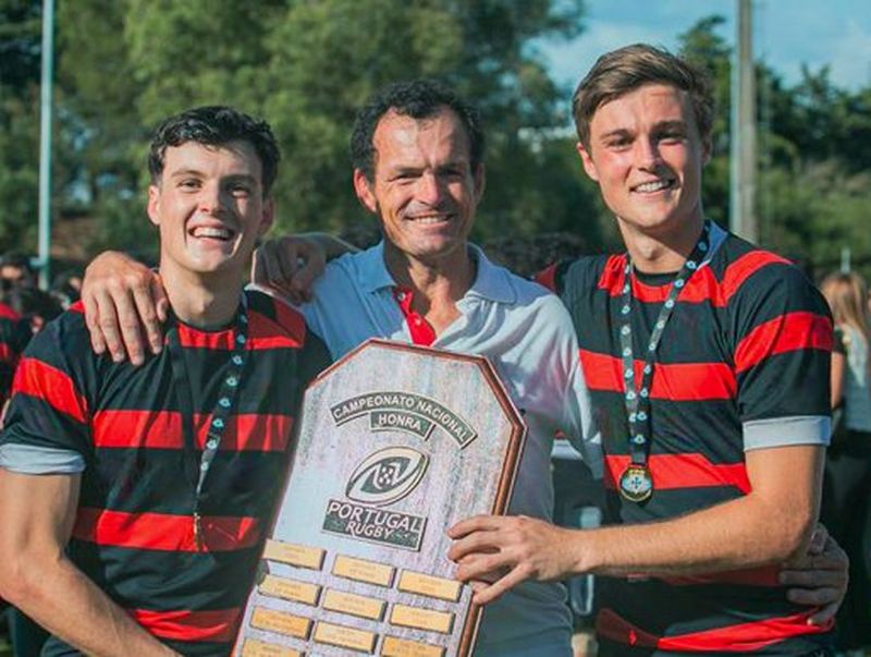 Jerónimo Portela (right), standing next to his father (centre) and a team-mate, wearing red-and-black rugby jerseys while holding a trophy