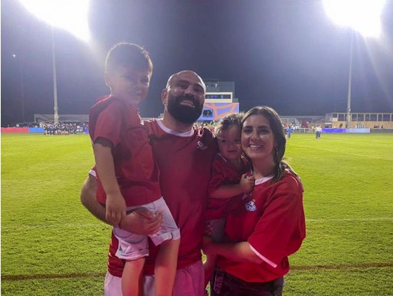 Mike Tadjer stands next to a floodlit rugby pitch wearing a Portugal rugby jersey, standing next to his wife, and holding his young sons.