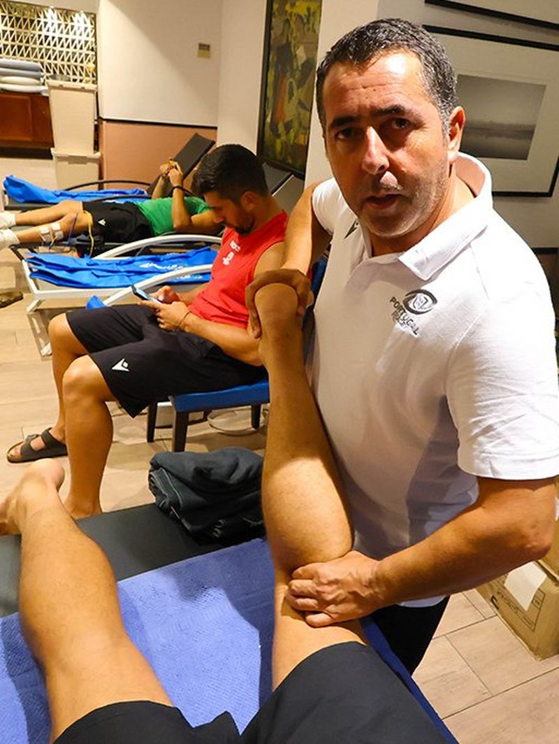 José Rodrigues massages a player’s left leg in the treatment room while he looks into the camera