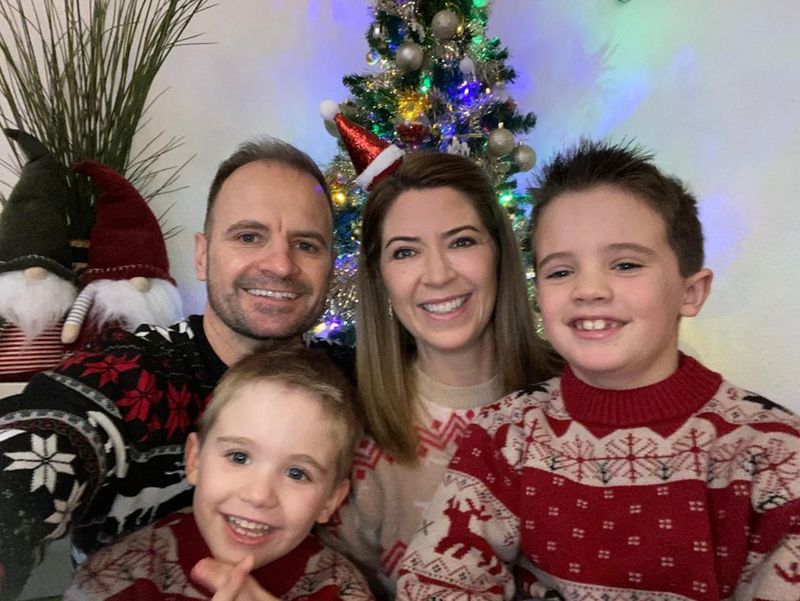 Sanette, her husband and two children pose for the camera, wearing sweaters of a Christmas design. There is a Christmas tree behind them.