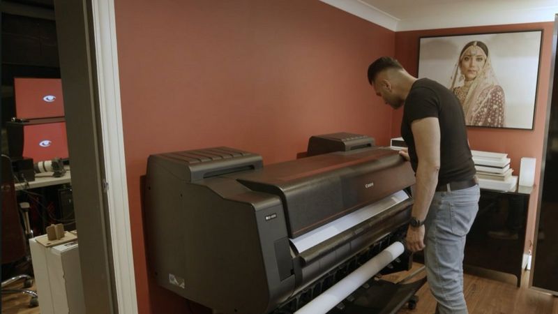 A man stood in a studio looking closely at large format printer, with framed portrait image on the wall in the background.