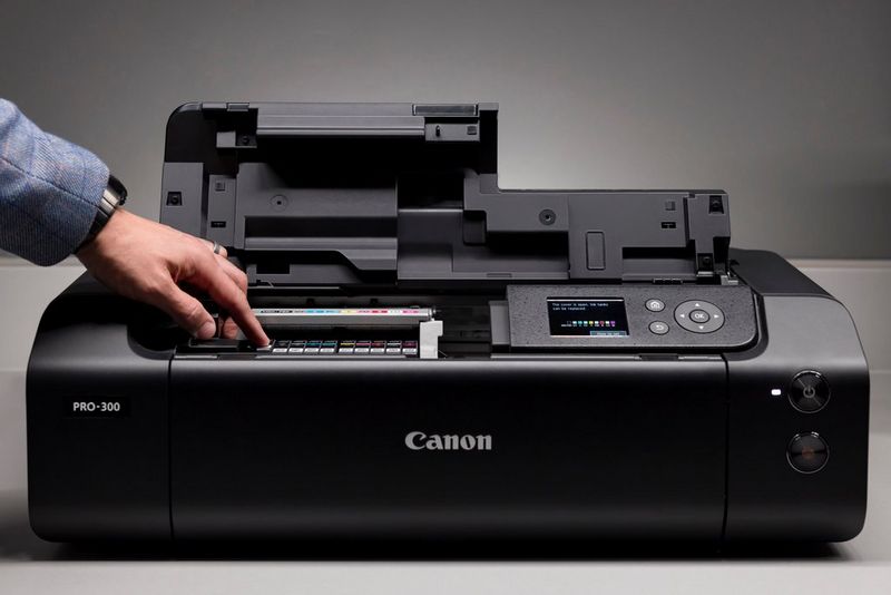 A Canon imagePROGRAF PRO-300 printer with its top cover open and a hand pressing one of the 10 ink cartridges into place. 