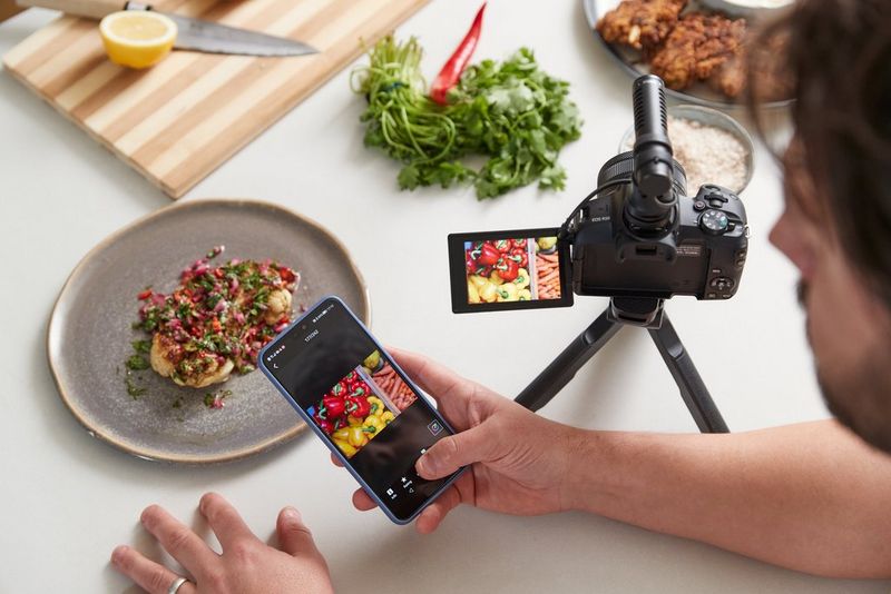 Talíř s jídlem je položen na stole vedle fotoaparátu Canon EOS R50 na stativu. Osoba drží v ruce chytrý telefon a na displeji je zobrazen stejný snímek jako na obrazovce fotoaparátu.