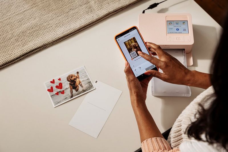 אישה משתמשת באפליקציה SELPHY Photo Layout של Canon בטלפון חכם כדי להוסיף קוד QR לתמונה, עם מדפסת SELPHY CP1500 על שולחן ברקע.