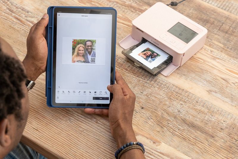 Ruke drže tablet s prikazanom slikom nasmiješenog para s QR kodom u kutu i opcijama uređivanja u aplikaciji SELPHY Photo Layout.