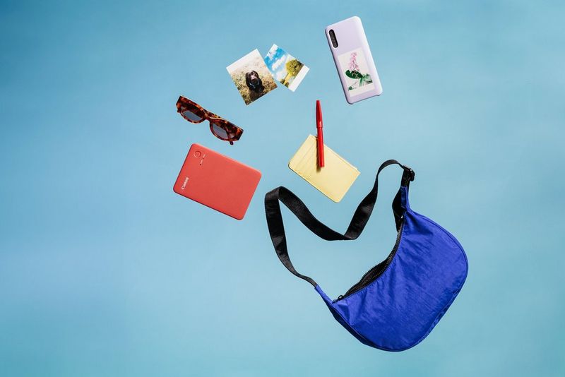 A Canon SELPHY QX20 printer is as if flying out of a small bag, surrounded by accessories. 