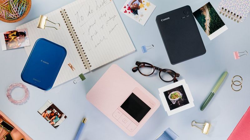 A pink Canon SELPHY CP1500, a black Canon SELPHY QX20 and blue Canon Zoemini 2 are spread out, surrounded by prints, stickers and accessories.