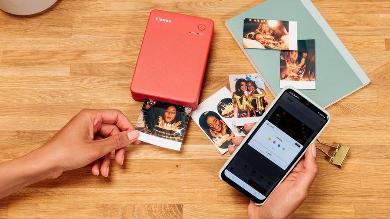 Een rode draagbare Canon-printer op een houten tafel produceert een kleine print zonder witrand van een vrouw die kaarsen op een verjaardagstaart uitblaast. De gebruiker houdt een smartphone vast waarop de Canon Mini Print-app wordt weergegeven en er liggen verschillende andere kleine fotoprints op de tafel.