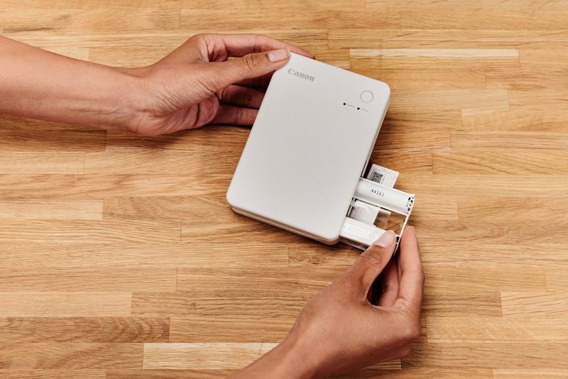 A pair of hands slot in the ink cartridges to the side of a Canon SELPHY QX20 printer.