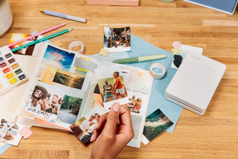 Рака држи фотографија на која има двајца луѓе и преклопена рамка. На масата во заднина има уште отпечатени фотографии, колаж, печатачот Canon SELPHY QX20 и канцелариски прибор.