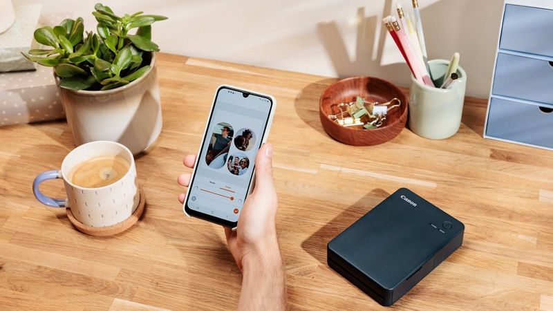 A hand holds a mobile phone displaying the Canon SELPHY Photo Layout App. On the table is a Canon SELPHY QX20, a cup of coffee and other stationery items.