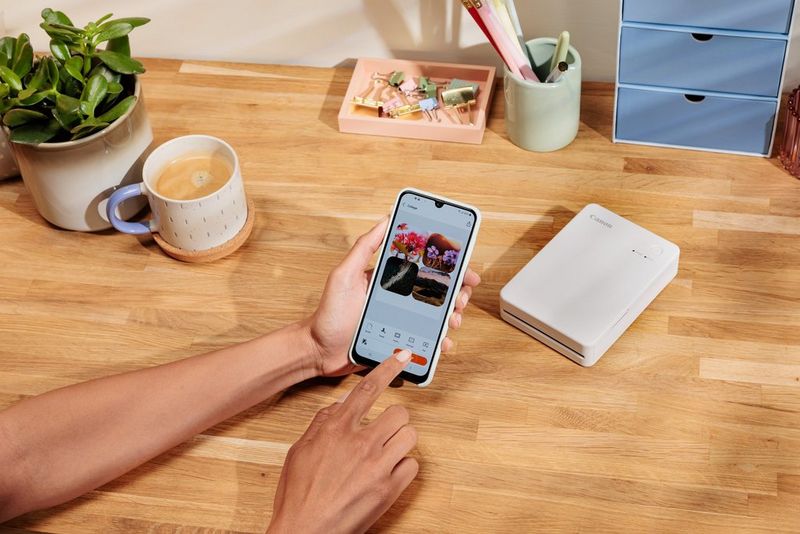Рака избира распоред на фотографии во апликацијата Canon SELPHY Photo Layout на паметен телефон, додека печатачот Canon SELPHY QX20 е поставен на масата во заднина. На масата има и шолја кафе, растение и канцелариски прибор.