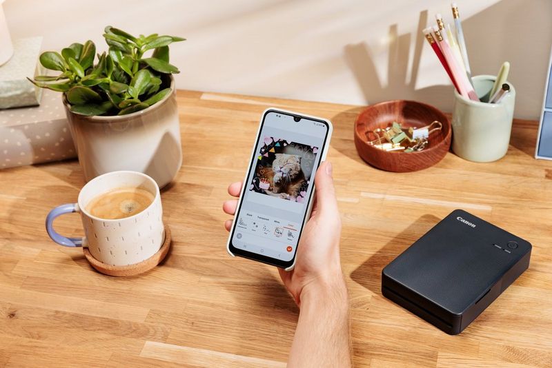 טלפון חכם עם תמונה של חתול עם קישוטים ועוגת יום הולדת באפליקציית SELPHY Photo Layout. Canon SELPHY QX20 שחורה על השולחן ברקע.