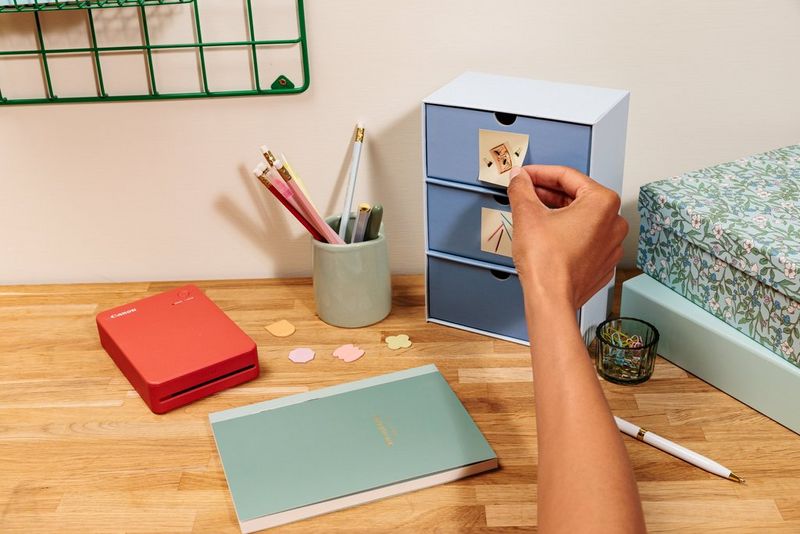 A user's hand attaches a sticker to the top drawer of a small stack of three on a desk with some craft paper, decorated boxes, pens and pencils in a jar, and a Canon SELPHY QX20 printer.