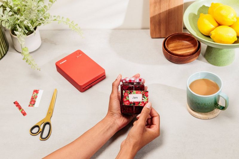A user applies a colourful picture sticker to a jar of strawberry jam. On the desktop behind are a cup of tea, a pair of scissors and a Canon SELPHY QX20 portable printer.