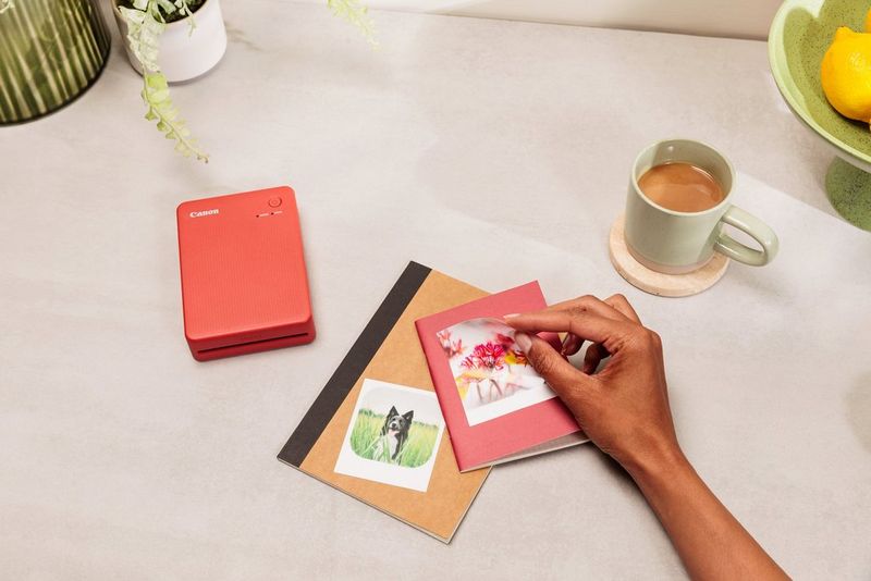A hand applies a square sticker to the smaller of two notebooks on a table, with a cup of tea to one side and a Canon SELPHY QX20 printer to the other.