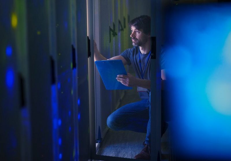 Blue-tinged view of a man in jeans and T-shirt referring to a clipboard while checking an IT data centre.