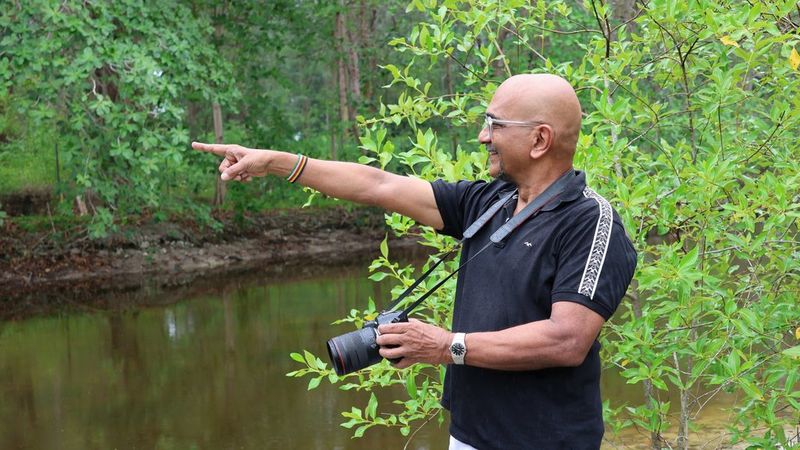 Dr Shah stands by a body of water and points with his right hand. In his left is a Canon camera, poised and ready to lift.