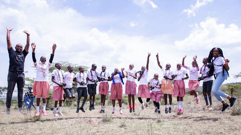 Sixteen people in a line, all jumping in the air. At one end is Canon Certified Trainer, Ben Mwangi, dressed all in black, and at the other is Wanjiru Ndegwa, in jeans and a white t-shirt. The children in between are wearing white t-shirts and either back shorts or red and white checked skirts. Some are holding cameras.