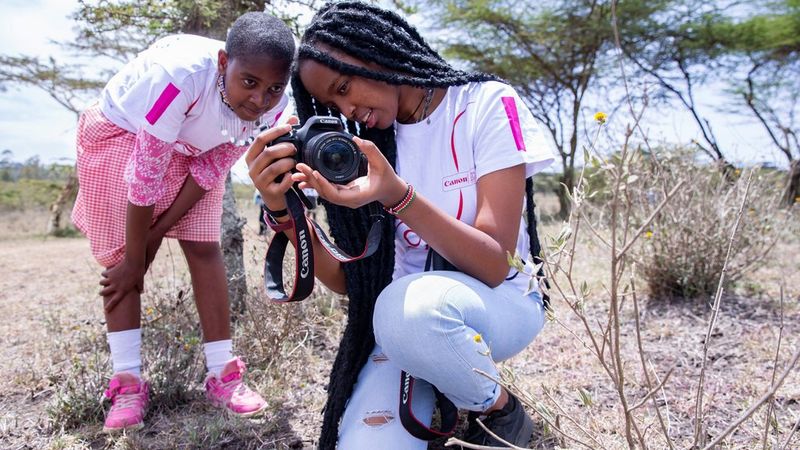 On the right, Wanjiru Ndegwa kneels with one knee on the ground. In her hand is a camera, of which she is showing the viewfinder to another child. Wanjiru wears a white and red Canon branded t-shirt and jeans. Her companion also wears a white and red Canon branded t-shirt, but with a red and white checked skirt, white socks and pink trainers.