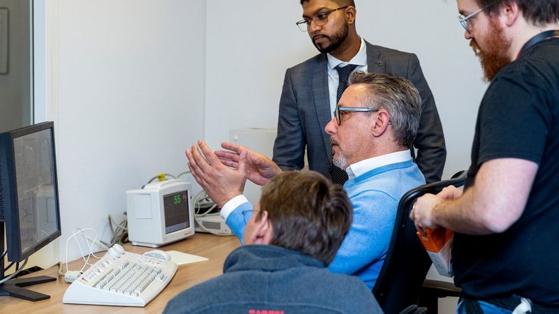 A man sits at a computer, gesturing to the screen. He is accompanied by three other men, who are also looking at his screen and listening.