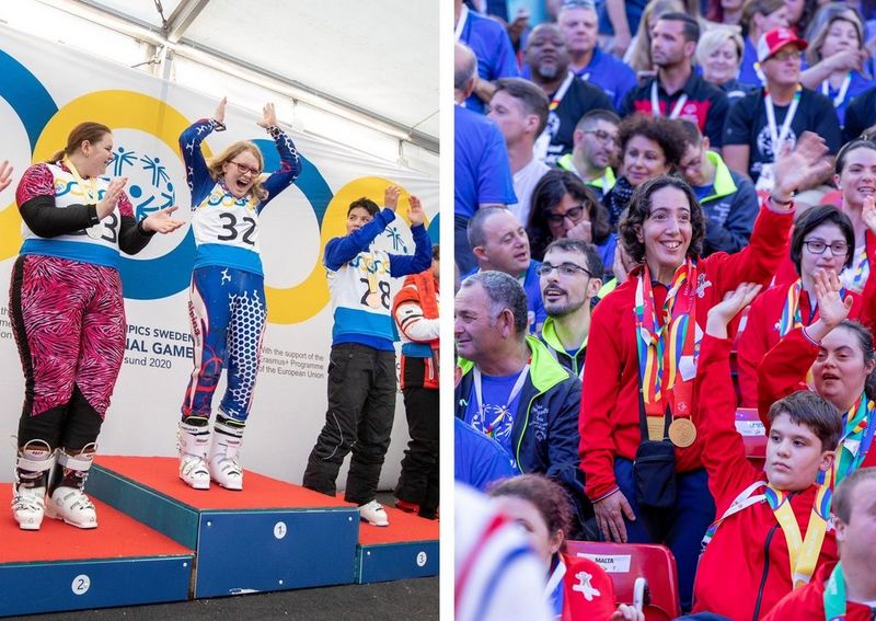 Three athletes on the podium. In the silver position, left, a woman in black and pink tight-fitting ski suit wears a competitor’s bib, her medal and ski boots. She is about to clap her hands. In the gold spot, the athlete wears a ski suit patterned in blue, red and white. Her bib bears the number 32, and she has her arms raised above her head with joy. In the bronze position, right, the athlete is of indeterminate gender and wears black trousers with a blue top and a bib bearing the number 28, but a medal obscures it. Behind them is a banner showing four blue and yellow rings and snippets of text about the event.