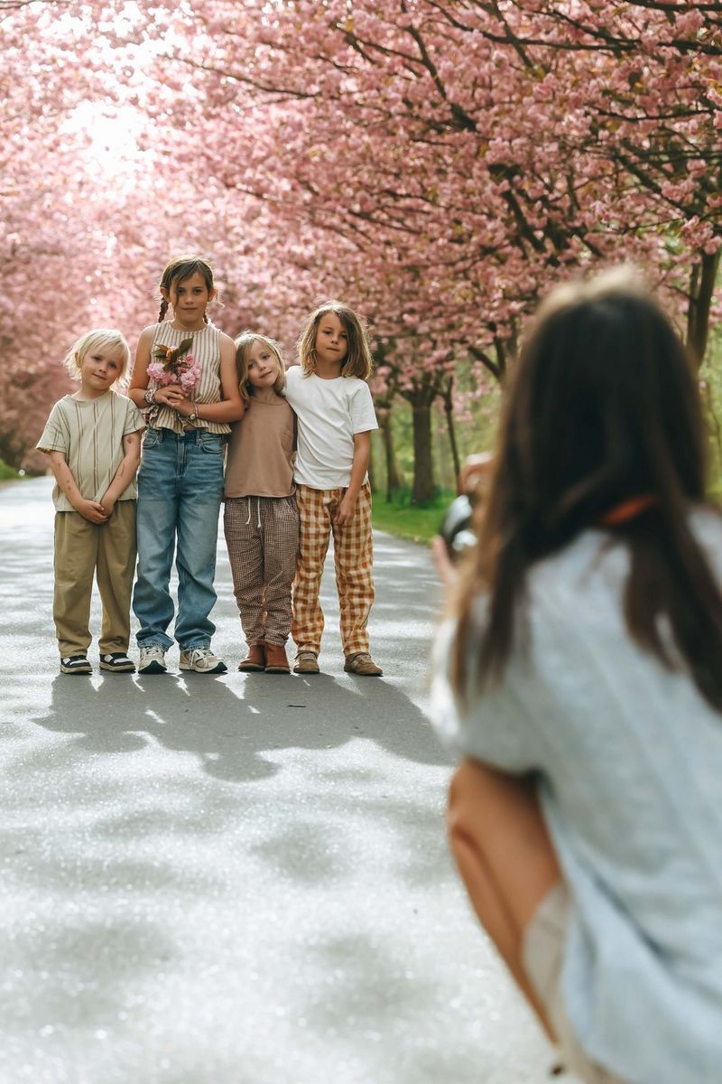 Four children stand together on a tree-lined path beneath blooming cherry trees while a photographer captures their portrait in the foreground.