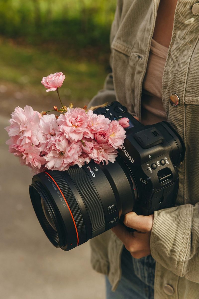 A close-up of a Canon EOS R5 Mark II camera fitted with a professional lens, held at waist level outdoors. Pink spring blossoms rest gently across the top of the lens, with soft natural light and a blurred green background creating a calm, seasonal atmosphere.